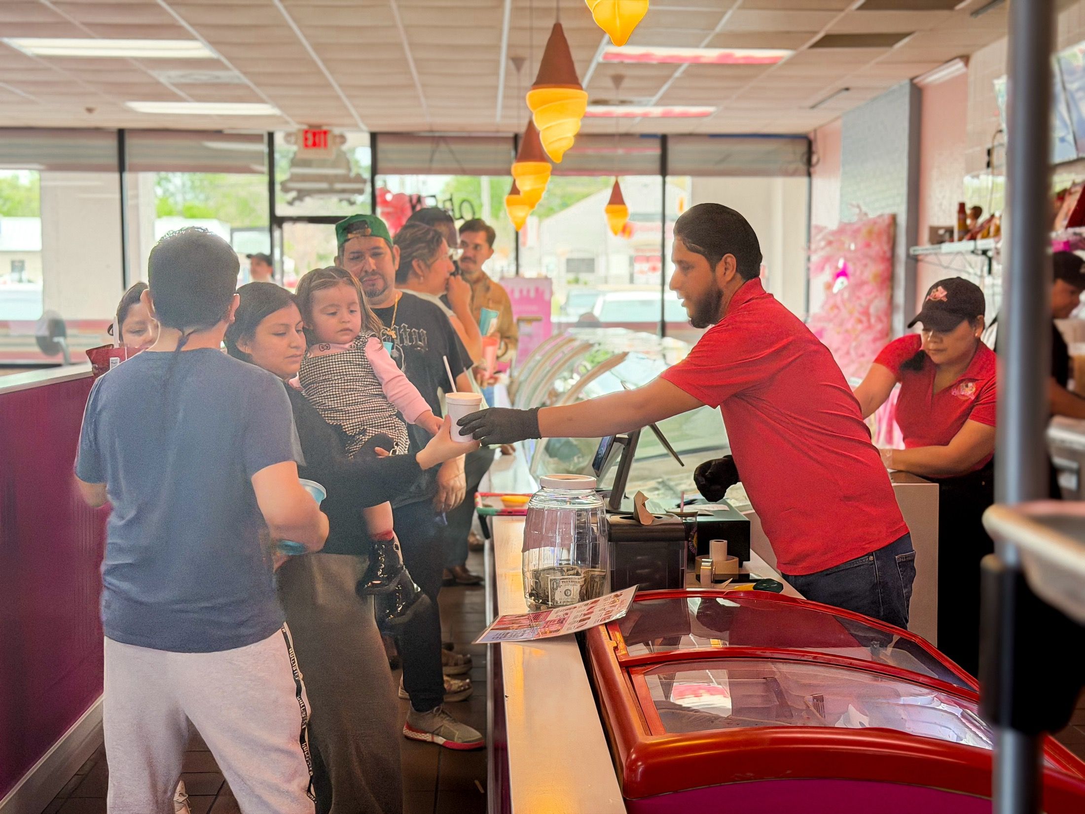 Families lined up at the Las Delicias counter under ice cream cone lights while a team member hands a cup across